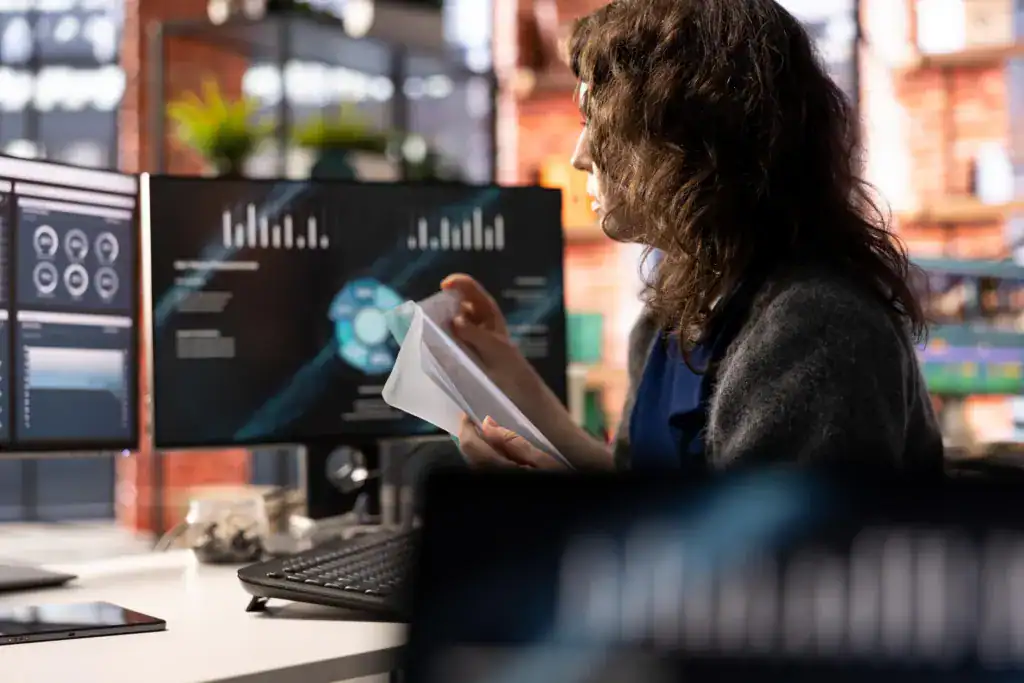 Woman reading a printed report beside monitors showing business dashboards and charts