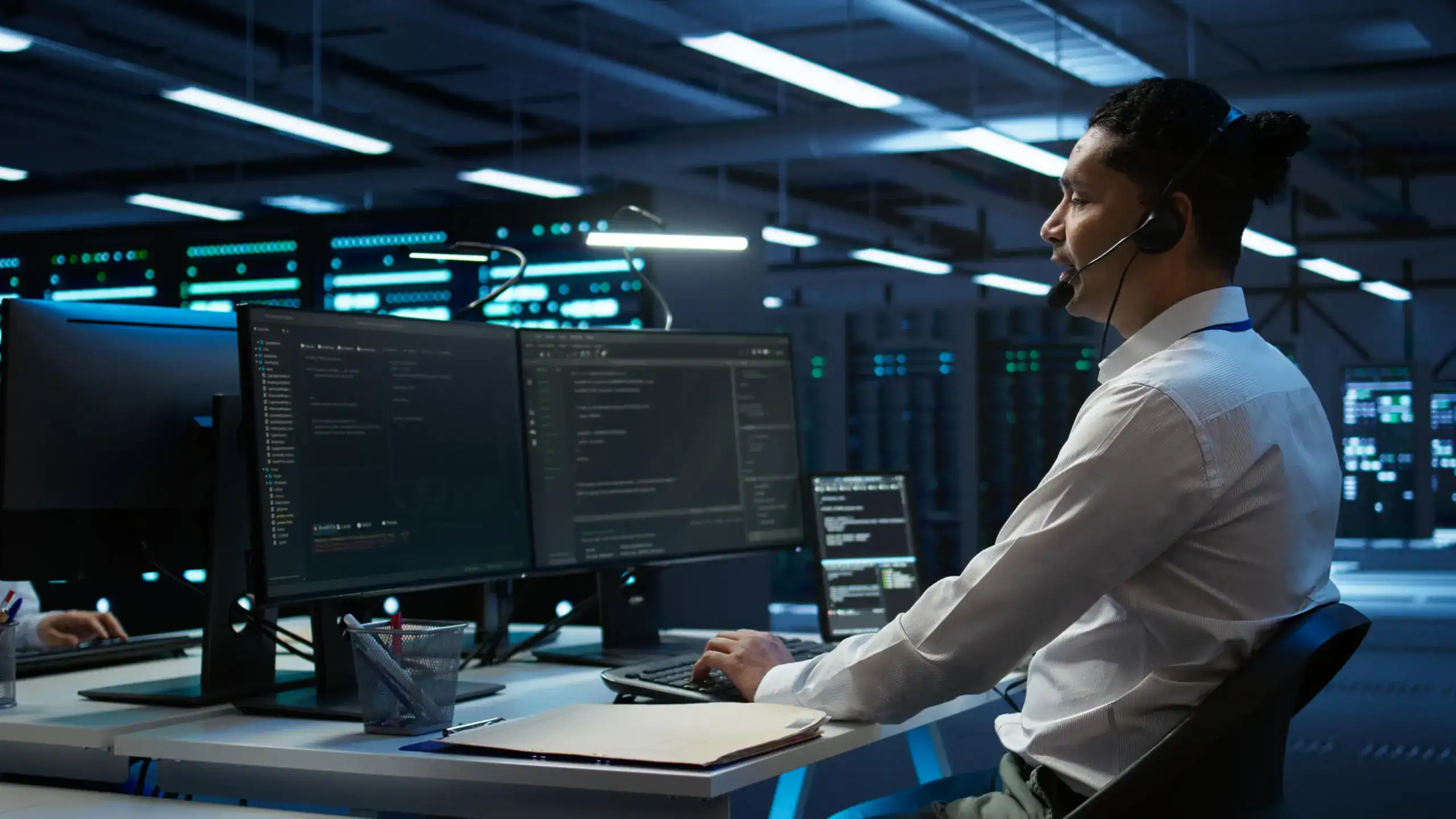 IT professional in a headset working across dual monitors of code inside a data centre