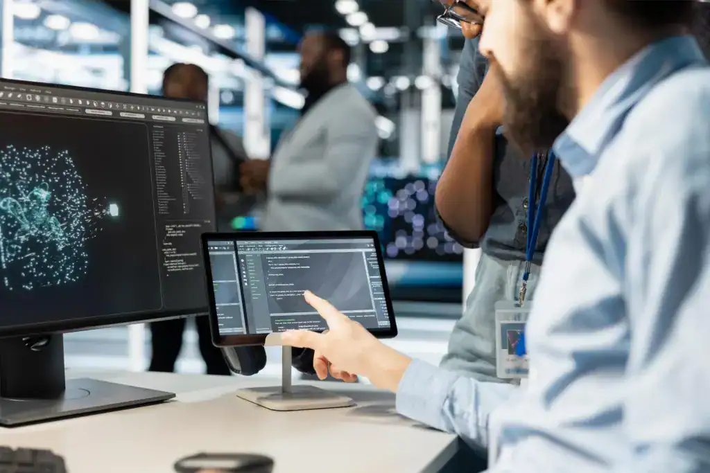 Two colleagues reviewing application code on a tablet and monitor in a technology office
