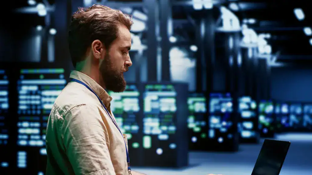 Technician using a laptop in a data centre, with racks of servers and green status lights glowing behind them